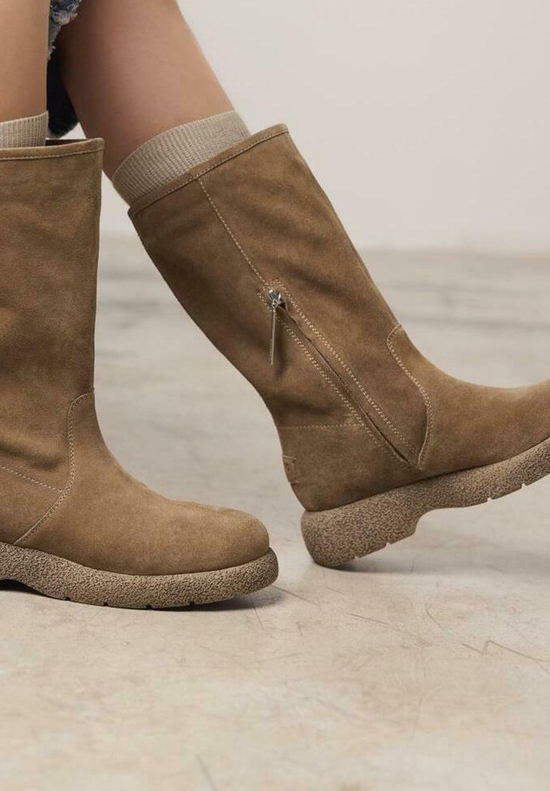 Botas altas de ante marrón con suela texturizada, cremallera lateral y puño de punto en la parte superior, exhibidas sobre una superficie de concreto.