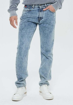 Personne portant un jean délavé bleu clair avec les mains près des poches et des baskets blanches sur fond uni.