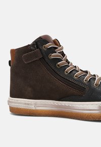 Baskets montantes en daim marron et panneaux en cuir noir, lacets beige, fermeture éclair latérale, bride Velcro, et semelle blanche en caoutchouc avec semelle extérieure en gomme.