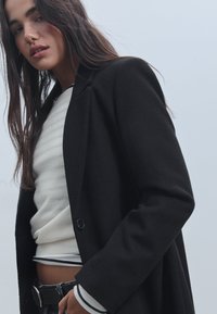 Mustavillainen villakangastakki, jossa on yksi nappi, puettuna kermanvärisen raidallisen neuleen päälle. Takissa on muotoiltu malli ja sileä tekstuuri.