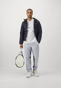 Μπουφάν παραγεμισμένο σε ναυτικό μπλε με μαύρη κουκούλα, λευκό πόλο πουκάμισο, ανοιχτό μπλε φόρμα με σκουρόχρωμες ρίγες στο πλάι, κρατάει ρακέτα τένις, λευκά αθλητικά παπούτσια.