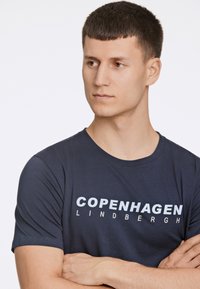 Tummansininen lyhythihainen t-paita, joka on valmistettu pehmeästä kankaasta ja jossa on rohkeaa valkoista tekstiä "COPENHAGEN LINDBERGH" edessä. Yksinkertainen muotoilu.