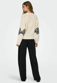 Crèmewitte gebreide trui met geribbelde structuur en contrasterende zwarte geometrische patronen op de mouwen, gecombineerd met zwarte broek met wijde pijpen.