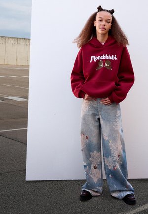 Jonge vrouw met krullend haar draagt een rode Monchhichi-hoodie en wijde jeans met verfspetters, staande tegen een witte muur buiten.
