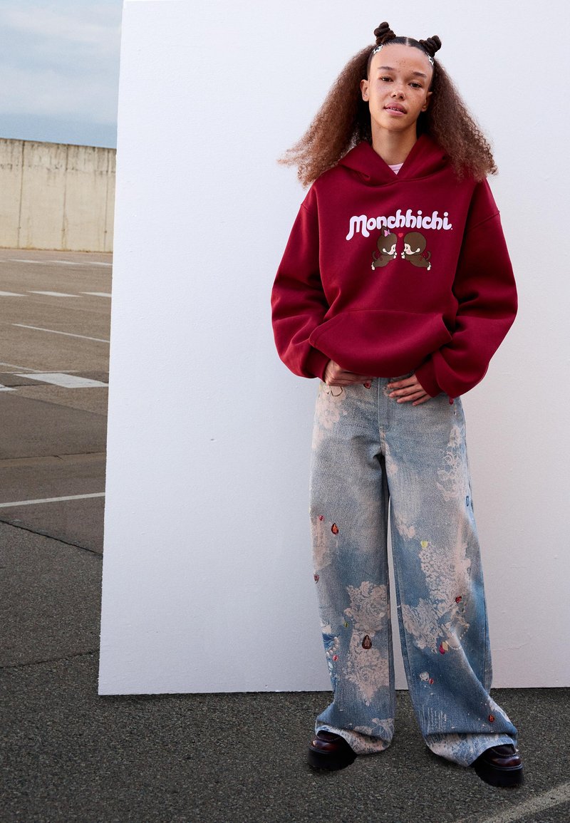 Giovane donna con capelli ricci indossa un hoodie Monchhichi rosso e jeans a gamba larga macchiati di vernice, in piedi contro un muro bianco all'aperto.