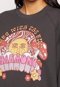Sudadera gris oscuro con un gran gráfico colorido que presenta un sol, champiñones y flores. El diseño incluye un texto en negrita que dice "ARMONÍA."