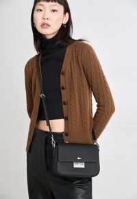 Cardigan en tricot brun, col roulé noir, pantalon en cuir noir et un petit sac noir avec des accessoires en argent, porté en bandoulière.