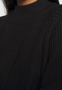 Schwarzer gestrickter Pullover mit geripptem hohen Kragen und strukturierten vertikalen Mustern auf der Brust und den Schultern.