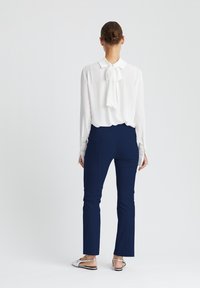 Witte blouse met een strik bij de hals, gecombineerd met marineblauwe aansluitende broek. Model draagt platte zilveren sandalen, gezien van achteren tegen een lichte achtergrond.