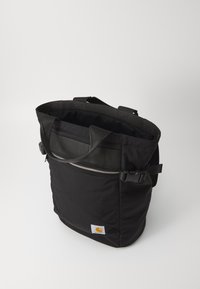 Sac fourre-tout en tissu noir avec doubles poignées, grande ouverture, poche zippée extérieure et patch logo. Matériau texturé, design simple.