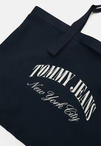 Sac fourre-tout en tissu bleu foncé avec "TOMMY JEANS New York City" imprimé en texte blanc incurvé à l'avant et deux poignées.