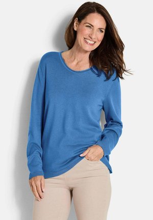 Lächelnde Frau mit langen braunen Haaren, die einen blauen Pullover mit langen Ärmeln und beige Hosen trägt, steht vor einem hellen Hintergrund.
