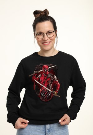 Smilende kvinde med briller iført en sort sweatshirt med Deadpool, der holder to sværd mod en mørk baggrund.