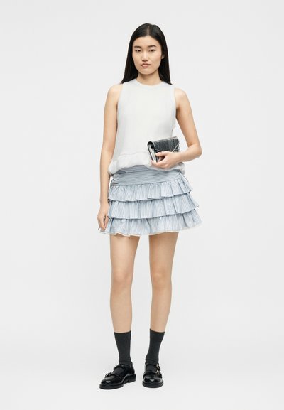 Femme portant un haut blanc sans manches et une mini-jupe à volants bleue à étages, tenant une pochette noire, avec des chaussettes noires et des mocassins noirs.