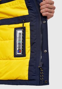 Intérieur de la veste isolante bleu marine et jaune. Comprend une doublure matelassée, une poche pour un thermomètre, une fermeture éclair noire et un cordon de serrage en accent.