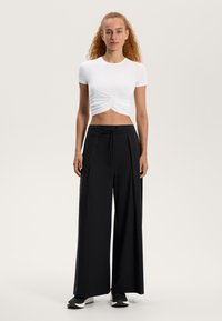 Valkoinen sovitettu lyhythihainen crop-top, jossa on kerrottu etuosa, yhdistetty korkeavyötäröisiin mustiin leveälahkeisiin housuihin, jotka sisältävät nauhavyötärön.