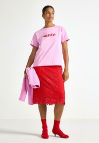 Lyserød bomulds T-shirt med "santé" trykt i rød, parret med en rød blonde-nederdel, lyserød strikket sjal og røde hælede sko med matchende strømper.