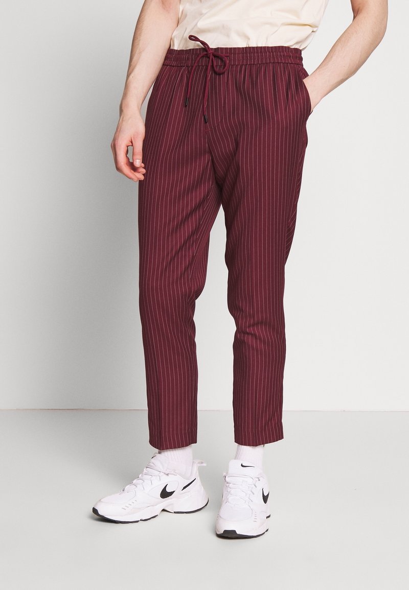 Homme portant un pantalon bordeaux à fines rayures avec cordon de serrage, des baskets blanches Nike et des chaussettes blanches, debout devant un fond clair uni.