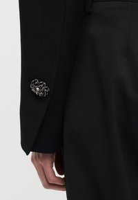 Μανίκι μαύρου κοστουμιού με περίτεχνο ασημένιο μανικετόκουμπο με λουλούδι και μερικώς ορατό χέρι σε ουδέτερο φόντο.