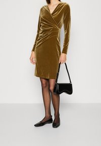 Robe en velours vert olive à manches longues et à design cache-cœur. Associée à des ballerines noires et un petit sac à main noir.