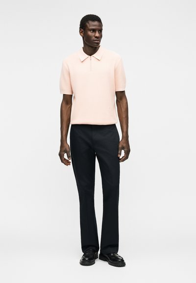 Άνδρας που φοράει ανοιχτό ροζ κοντομάνικο πουκάμισο με φερμουάρ στον γιακά, μαύρο παντελόνι και μαύρα παπούτσια, στέκεται μπροστά σε απλό φόντο.