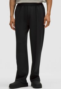 Zwarte, hoge taille broek met een gladde textuur, wijde pijpen en subtiele geplooide details aan de voorkant. Gekleed met zwarte instap schoenen.