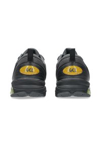 Zapatos deportivos negros con un upper sintético texturizado, lengüetas en el talón y un logo amarillo "GEL" en la parte trasera. Presenta un acento verde en la suela.