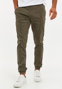 Uomo che indossa pantaloni jogger verde oliva e sneaker bianche, con le mani rilassate, in piedi contro uno sfondo bianco uniforme.