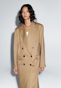 Brown double-breasted blazer dress with large lapels, button accents, and side pockets. Fabric has a smooth, slightly structured texture.
