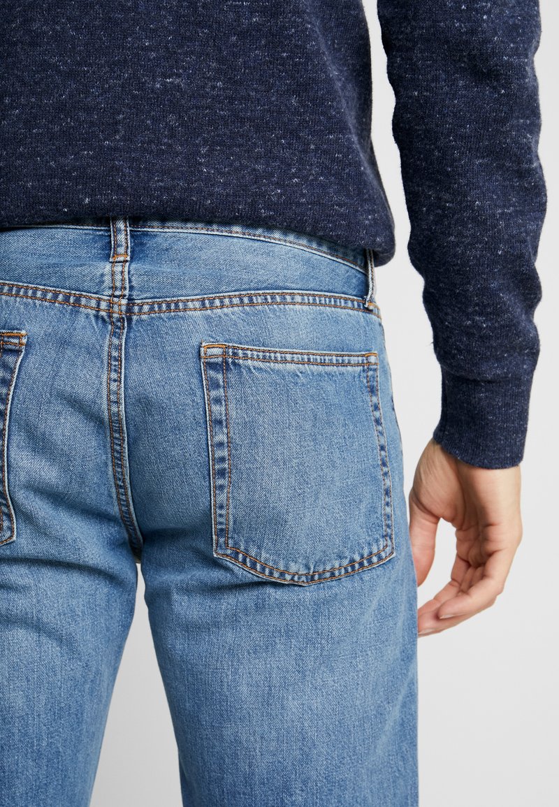 Helle blaue Jeans mit zwei Gesäßtaschen, sichtbaren Nähdetails und einer figurbetonten Passform, kombiniert mit einem dunkel strukturierten Pullover.
