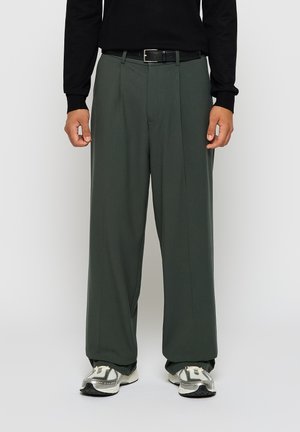 Loose, dark green trousers with pleats, made of smooth fabric. Paired with a black belt and gray sneakers, standing against a neutral backdrop.