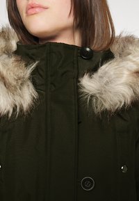 Parka vert olive avec une garniture en fourrure synthétique sur la capuche, dotée de boutons noirs, d'un col haut et de poches plaquées à l'avant. Texture douce visible.