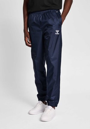 Marineblaue wasserdichte Jogginghose mit elastischem Bund und Bündchen. Merkmale ein weißes Logo auf dem linken Oberschenkel. Glattes, glänzendes Material.