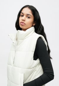 Φουσκωτό λευκό γιλέκο μ�ε ψηλό γιακά και φερμουάρ, που διαθέτει διακριτικό λογότυπο Nike στο στήθος και υφασμένη ραφή για επιπλέον λεπτομέρεια.