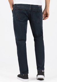 Dunkelblaue Jeans aus Denim mit schmaler Passform, Fünf-Taschen-Design und sichtbaren Nähten an den hinteren Taschen. Getragen mit schwarzen Sneakers.