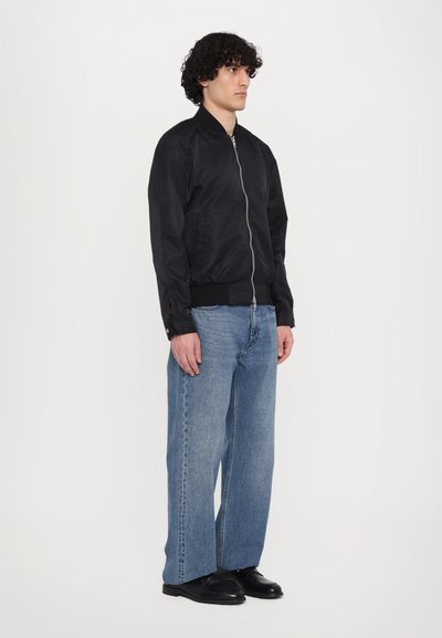 Zwarte bomberjack met een ritssluiting aan de voorkant, geribde manchetten en tailleband. Gepaard met lichtblauwe wijde jeans en zwarte schoenen.