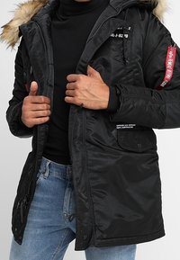 Parka noire avec capuche doublée de fourrure, devant zippé, de multiples poches, et des accents d'étiquette rouge, portée sur un col roulé noir.