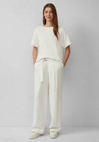 Biała koszulka z podwiniętymi rękawami, w zestawie z białymi spodniami z wysokim stanem i plisami oraz białymi trampkami, na tle jasnoszarym.