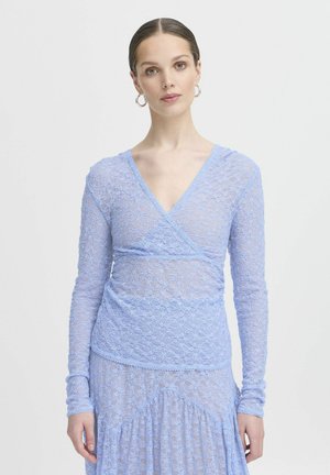 Top lungo a maniche con pizzo azzurro chiaro, presenta un design avvolgente, un intricato motivo floreale e bordi scallop. Il modello indossa una gonna abbinata.