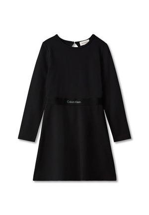 Junges Mädchen mit lockigem Haar, das ein schwarzes, langärmeliges Kleid und schwarze Loafers trägt und mit verschränkten Händen vor einem schlichten Hintergrund steht.