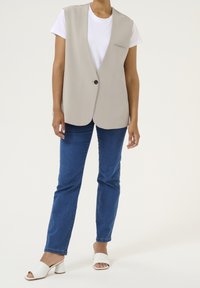 Beige ärmellose Weste mit einem Knopfverschluss und einer Brusttasche, kombiniert mit blauen Jeans und weißen High Heels.