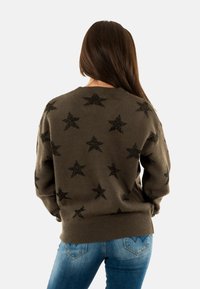 Donna con lunghi capelli castani, che indossa un maglione marrone con motivi a stelle nere e jeans blu, vista da dietro.
