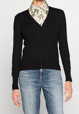 Personne portant un cardigan noir en maille torsadée boutonné sur une chemise à motifs boutonnée et un jean bleu délavé sur un fond neutre.