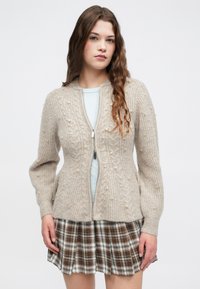 Jovem mulher com cabelo ondulado e longo, vestindo um cardigã de malha bege com detalhes em pérola sobre uma blusa azul e uma saia xadrez castanha, em pé contra um fundo branco.