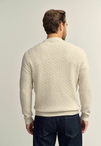 Maglione di colore crema, con texture e motivo a griglia, polsini e orlo a costine, indossato con jeans blu scuro, visto da dietro.