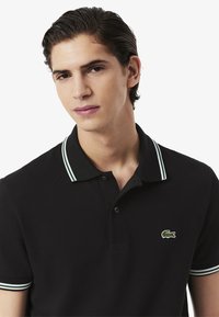 Svart pikétröja med grön och vit randig krage, texturerat tyg, tvåknappsslutning och en liten broderad logotyp på bröstet.