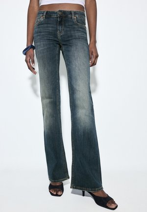 Frau trägt ausgewaschene blaue hoch taillierte Schlagjeans, kurzes weißes Oberteil, schwarze Sandalen mit Absatz und ein dunkelblaues Armband am linken Handgelenk.