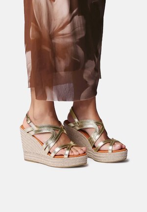 Pies con sandalias de cuña metálicas doradas con suelas de plataforma de yute trenzado, bajo una falda marrón con estampado floral y fluida.