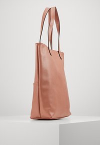 Růžová kožená taška přes rameno s plochou, hladkou texturou a dlouhými popruhy. Obsahuje boční kapsu a minimalistický design, umístěná na bílém podstavci.