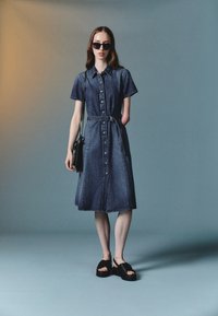 Denim shirt dress, short sleeves, button-down front, belted waist, knee-length. Matched with black platform shoes and a small bag.